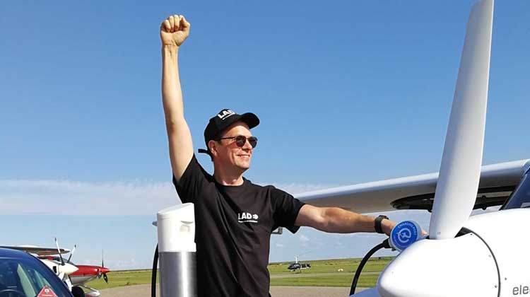 Dennis Schulmeyer mit Ladesäule an einem Elektroflugzeug. In der einen Hand hält er den Ladestecker an das Flugzeug, der andere Arm ist in die Höhe gestreckt.
