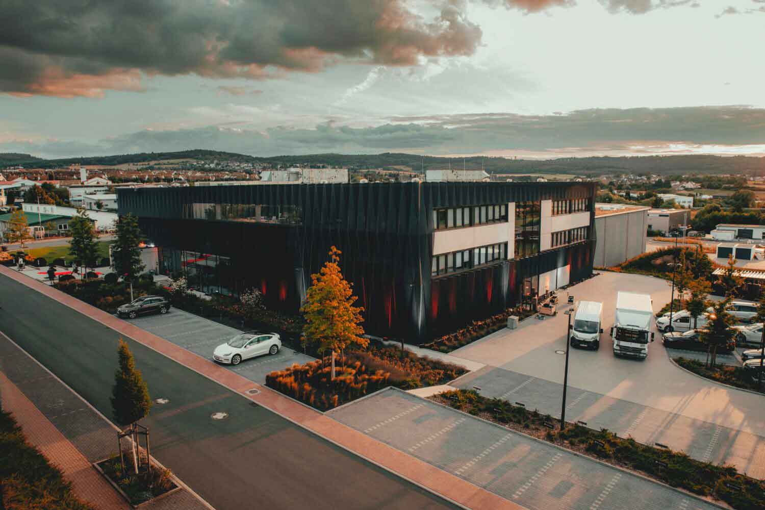 Foto von großem viereckigen Bürogebäude umgeben von vielen Parkplätzen auf denen zum Teil Elektrofahrzeuge stehen.
