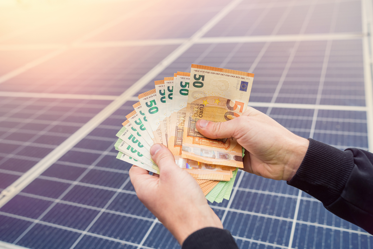 Zwei Hände halten viele 50-Euro-Scheine vor einer Photovoltaikanalage, auf die die Sonne scheint