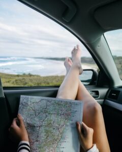 Person hält Beine aus dem Fahrerfenster eines Autos und hält dabei eine Landkarte in den Händen.